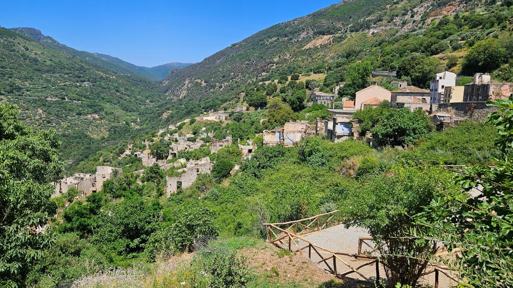 Blick auf ein verlassenes Dorf mit überwucherten Ruinen, umgeben von grünen Bergen und einem klaren blauen Himmel.