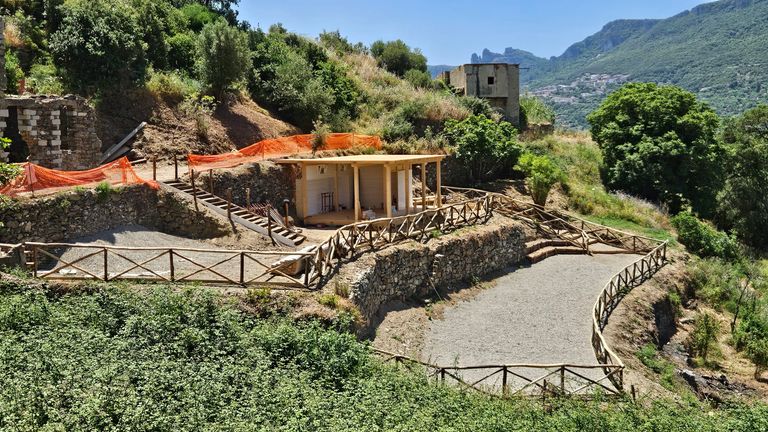 Eine antike Grabstätte mit überdachtem Altar und hölzernem Geländer, eingebettet in eine grüne Landschaft mit Hügeln und Bergen im Hintergrund.