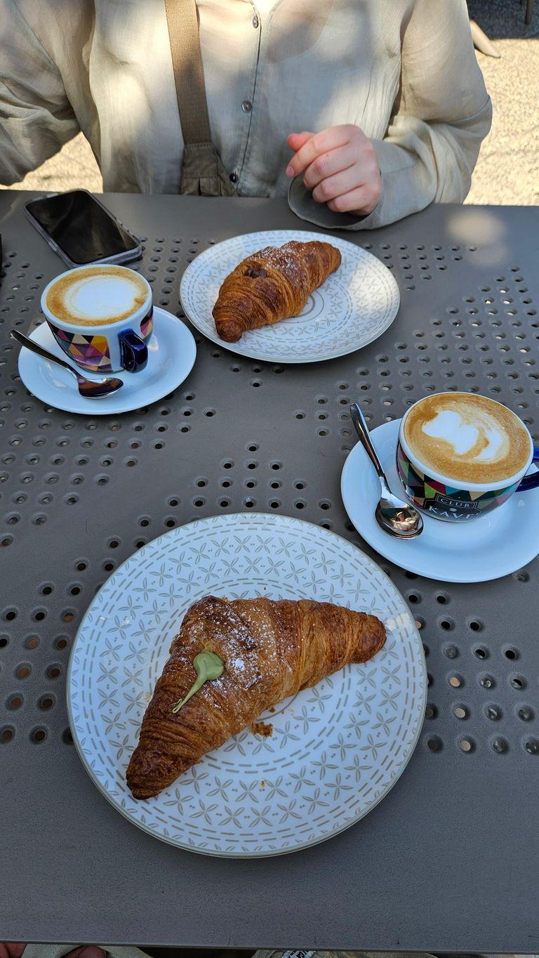 Zwei bunt gemusterte Kaffeetassen mit Löffeln auf Untertassen, jeweils mit Cappuccino gefüllt, stehen auf einem perforierten grauen Tisch. Zwei Croissants auf dekorativen weißen Tellern sind ebenfalls auf dem Tisch platziert, eine Person in heller Kleidung sitzt am Tisch.
