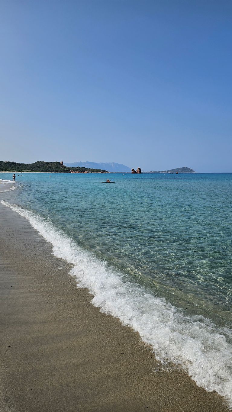 Sandstrand mit seichtem, klarem Wasser und kleinen Wellen unter blauem Himmel, im Hintergrund Personen im Wasser und weiter entfernte bewaldete Inseln.