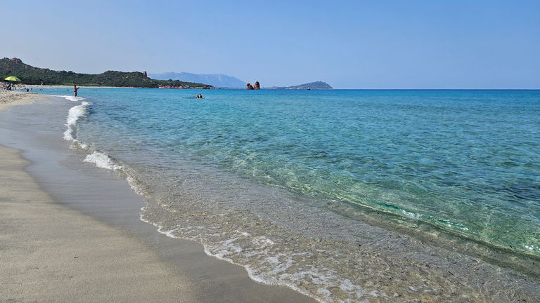 Sandstrand mit klarem, blauem Wasser und kleiner Brandung, im Hintergrund wenige Menschen und bewaldete Hügel unter blauem Himmel.
