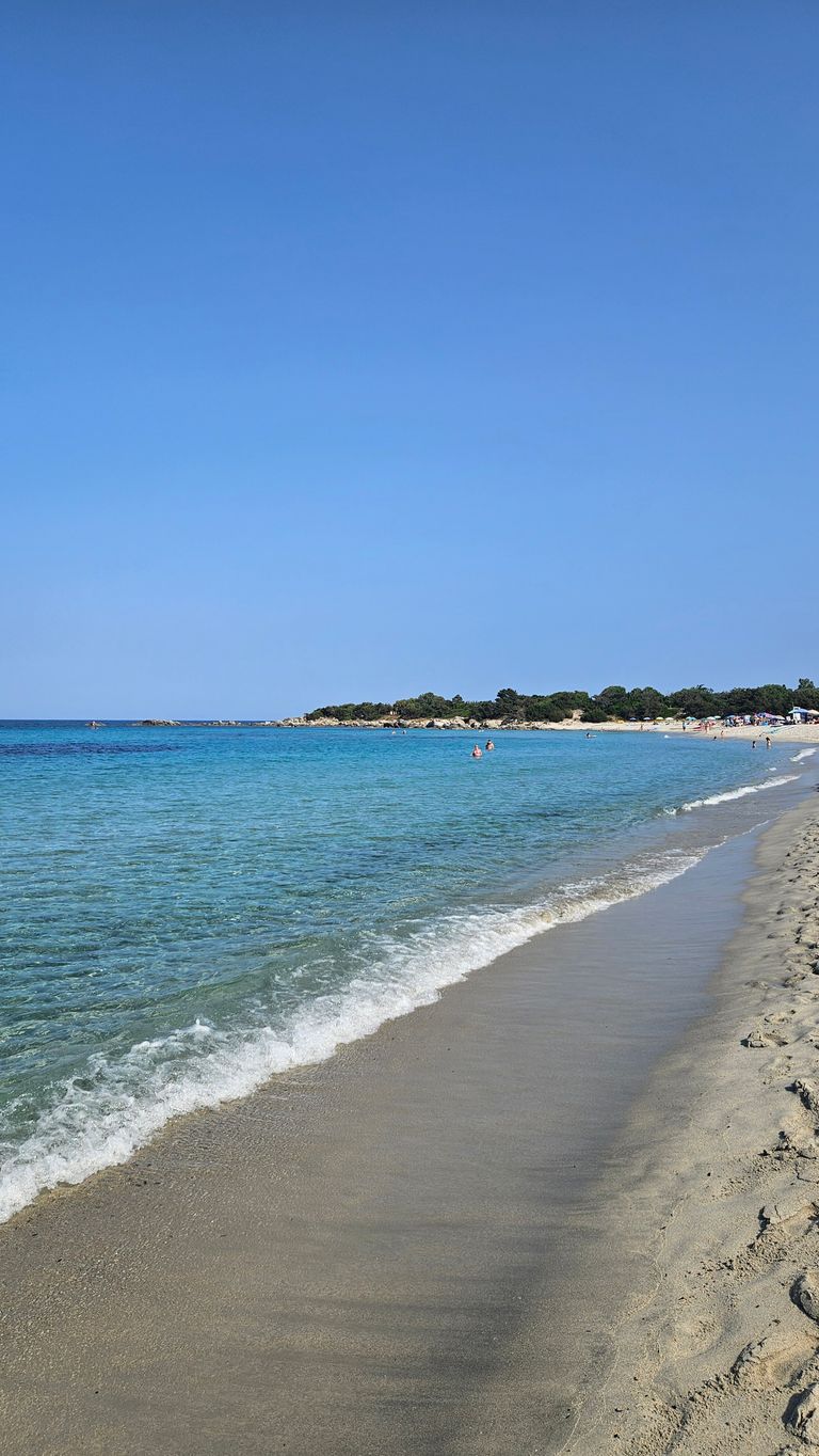 Ein weiter Sandstrand mit sanften Wellen, blauem Meer und klarem Himmel, am Horizont einige Menschen im Wasser und Bäume am Ufer.