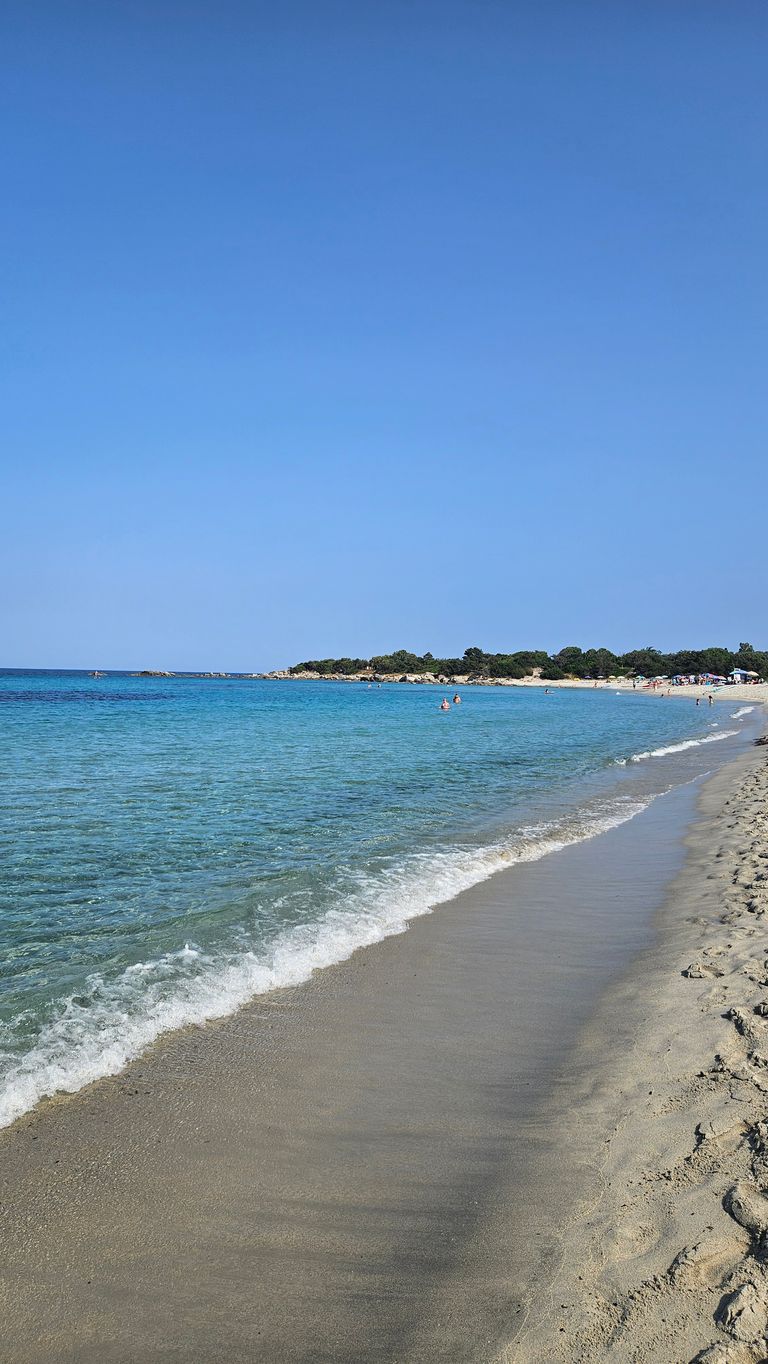 Sandstrand mit sanften Wellen und klar blauem Meer unter klarem Himmel, im Hintergrund Bäume und Menschen am Strand.