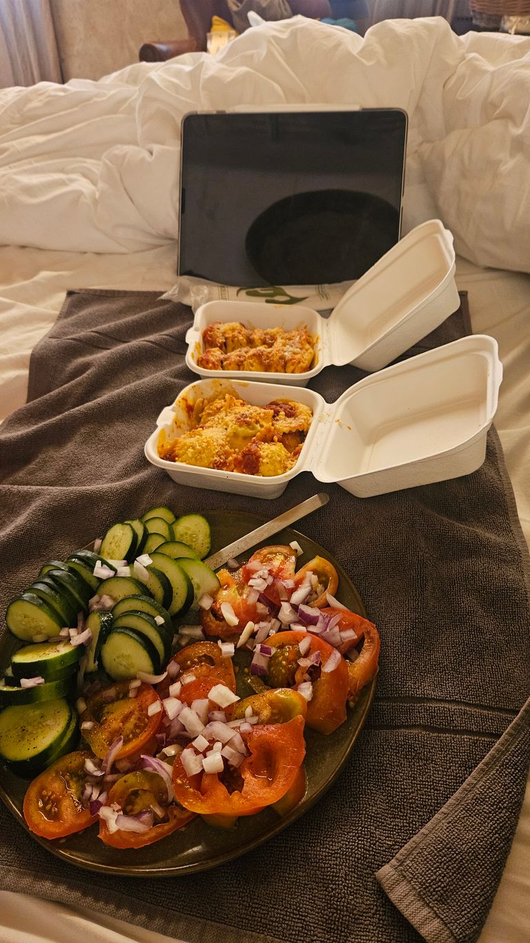 Zwei geöffnete weiße Pappbehälter mit zubereitetem Essen und ein Teller mit Tomaten, Gurkenscheiben und gehackten Zwiebeln auf einem braunen Handtuch auf einem Bett. Im Hintergrund steht ein Tablet vor einem ungemachten Bett.