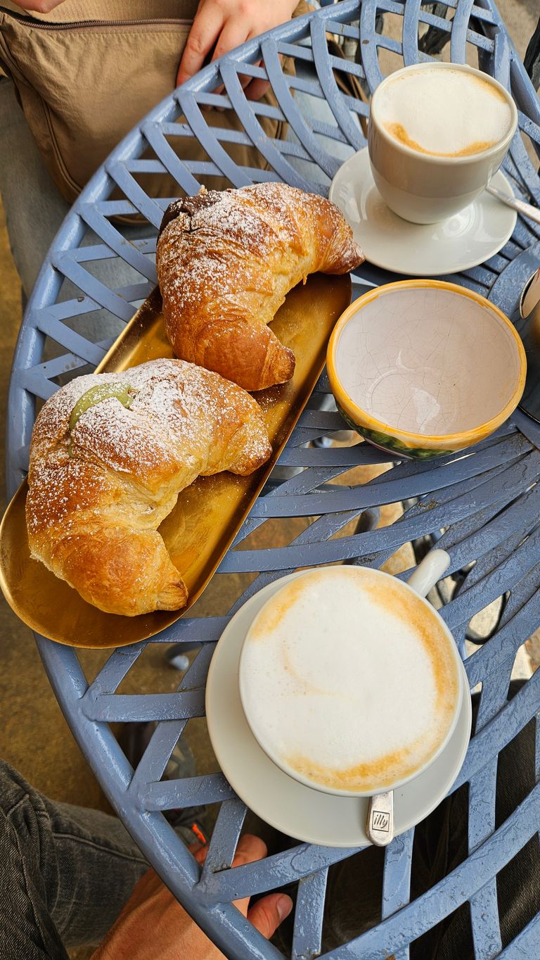Zwei Croissants mit Puderzucker auf einem goldfarbenen Tablett, dazu zwei Tassen Cappuccino auf einem blauen, metallenen Gartentisch.