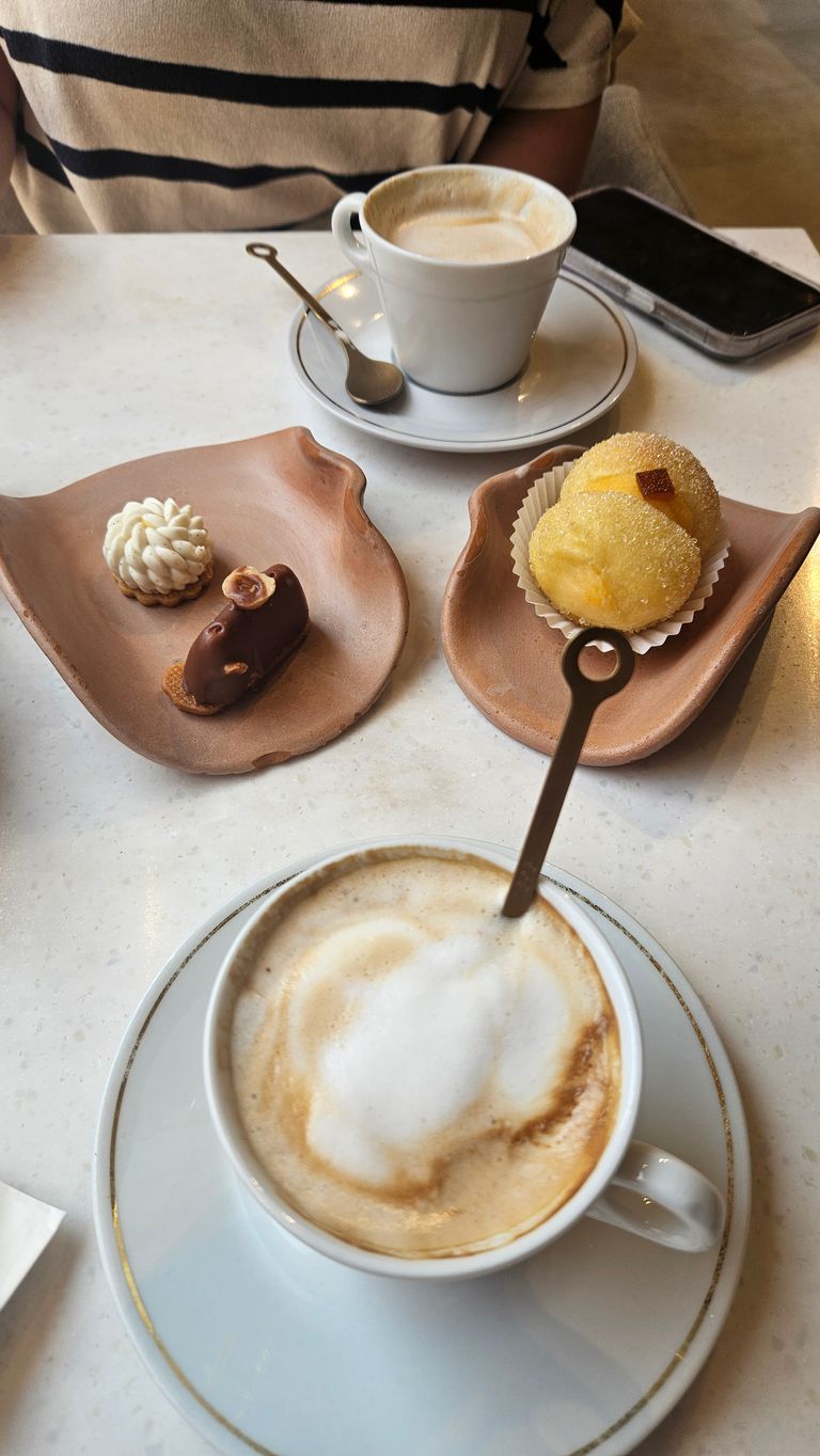 Zwei Tassen Kaffee auf einem weißen Tisch mit zwei kleinen Tellern, die verschiedene kleine Desserts enthalten, und einer Person mit gestreiftem Shirt im Hintergrund.