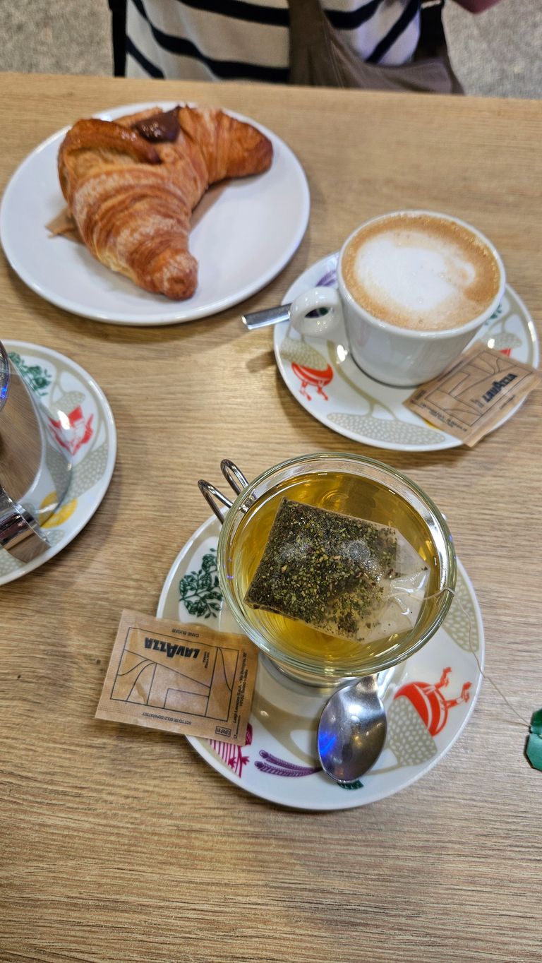 Auf einem Holztisch stehen eine Tasse Tee mit einem Teebeutel darin, eine Tasse Cappuccino und ein Teller mit einem Croissant. Auf den Untertellern liegen Zuckertütchen mit der Aufschrift Lavazza.