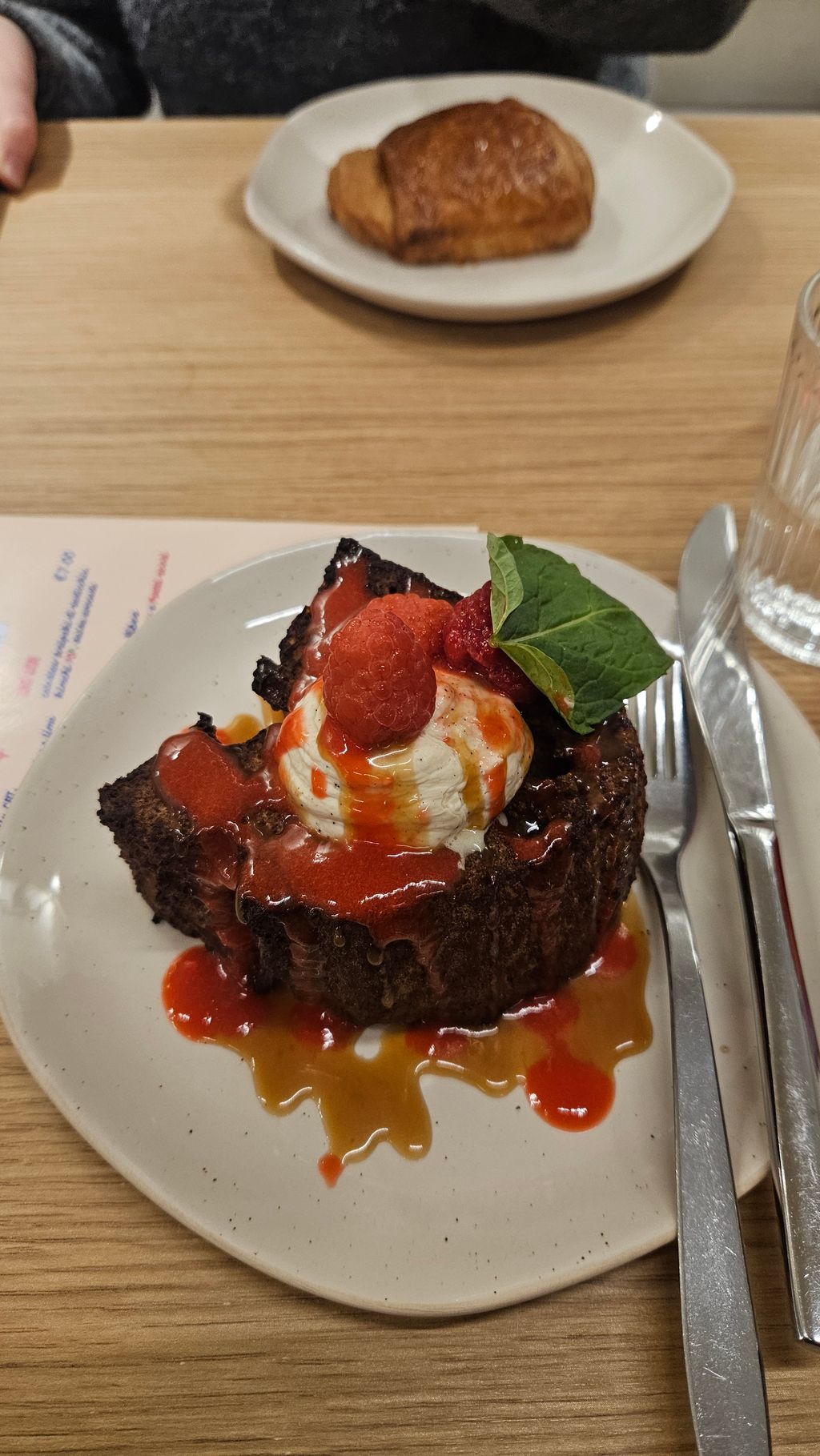 A piece of dark cake on a white plate topped with cream, raspberries, red sauce, and a green leaf. In the background, a brown pastry on another white plate on a wooden table.