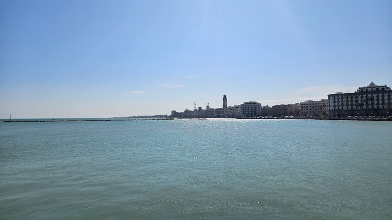 View of a city's waterfront along the sea under a clear blue sky with sunlight reflecting on the water surface.