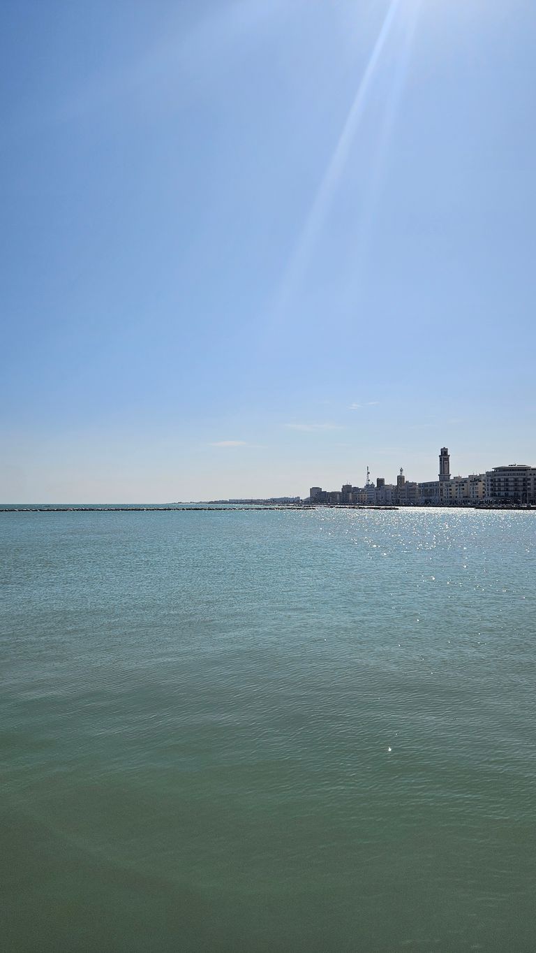 View of the sparkling sea with a waterfront promenade and buildings on the horizon under a clear blue sky with sun rays.