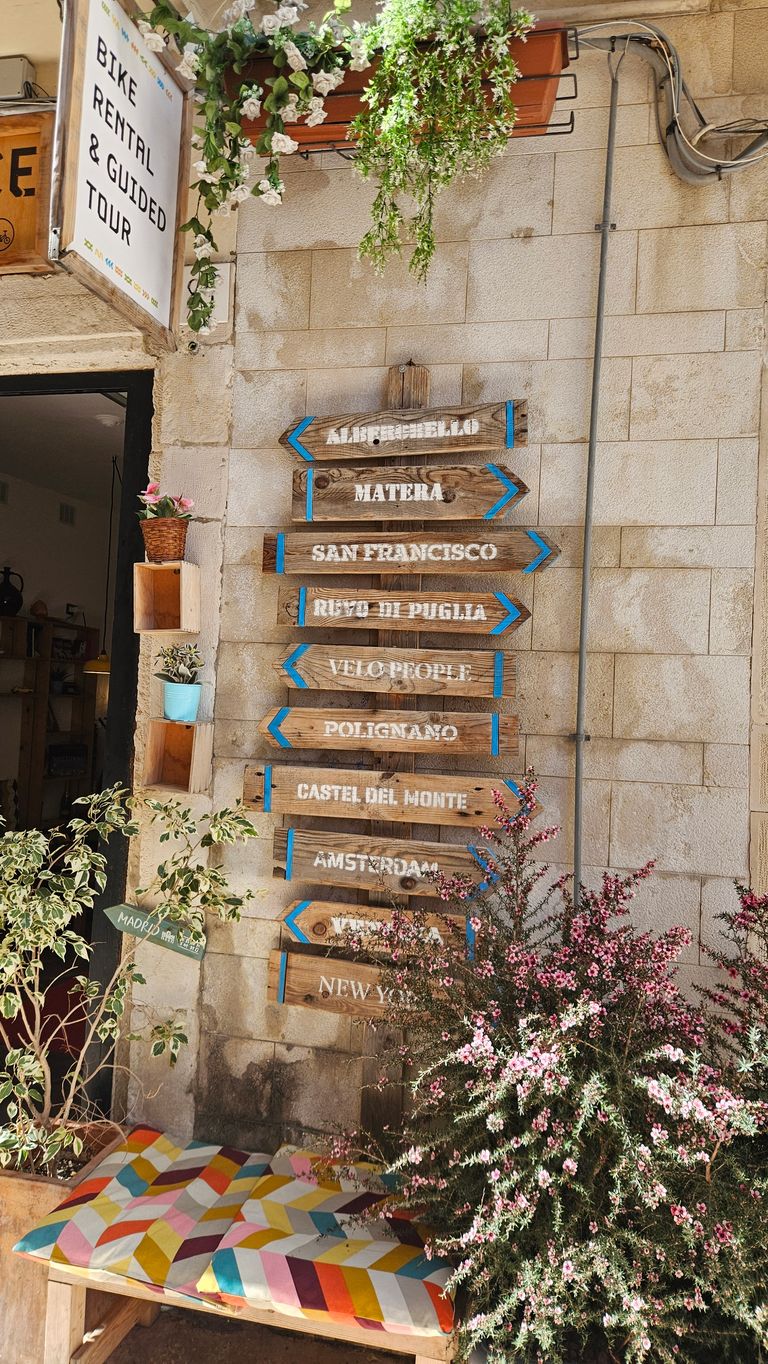Wooden directional signpost pointing to various cities, next to a wall adorned with flowers and a colorful cushion on a wooden bench.