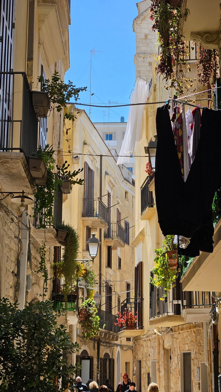 Narrow sunlit old town alley with closely built stone and stucco buildings, balcony plants, and clothes hanging out to dry.