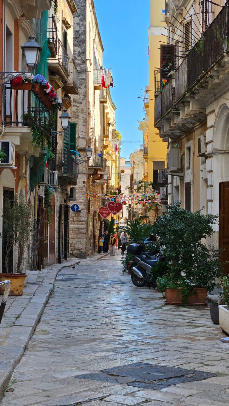 Narrow cobblestone street lined with old buildings, potted plants, and many street lamps, with people and colorful decorations including two heart-shaped signs with Italian text in the background.