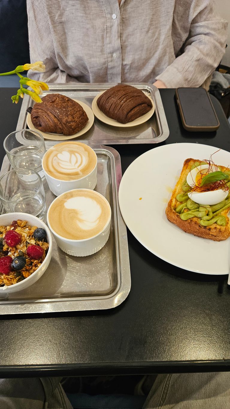 Breakfast table with two cappuccinos, granola with berries, two chocolate croissants, and an avocado toast topped with egg on a white plate. Person wearing a striped shirt in the background.