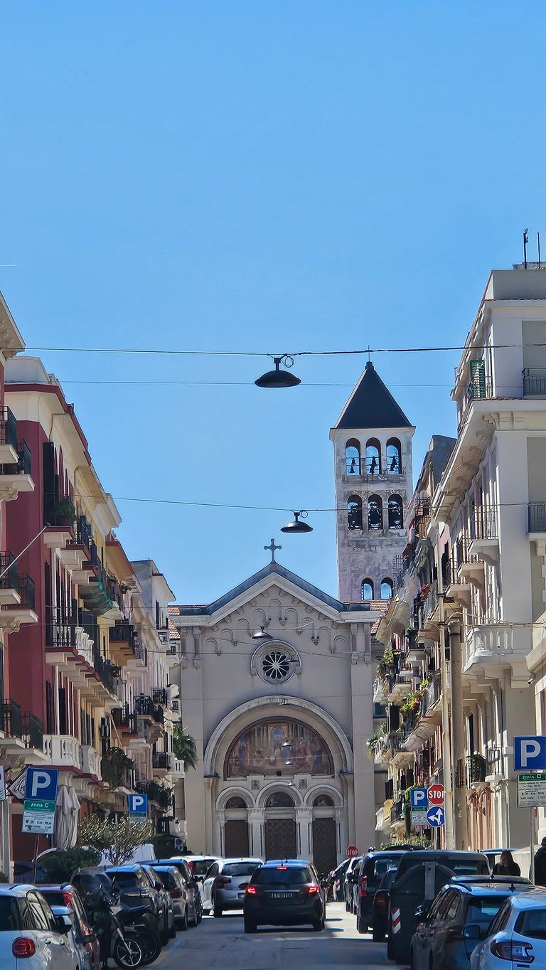 A narrow street view with parked cars and multi-story buildings on both sides, ending with a church featuring a bell tower under a clear blue sky.
