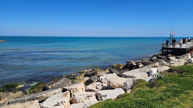 Rocks and green grass along the coast with clear blue sky and calm sea, people standing on a pier in the background.