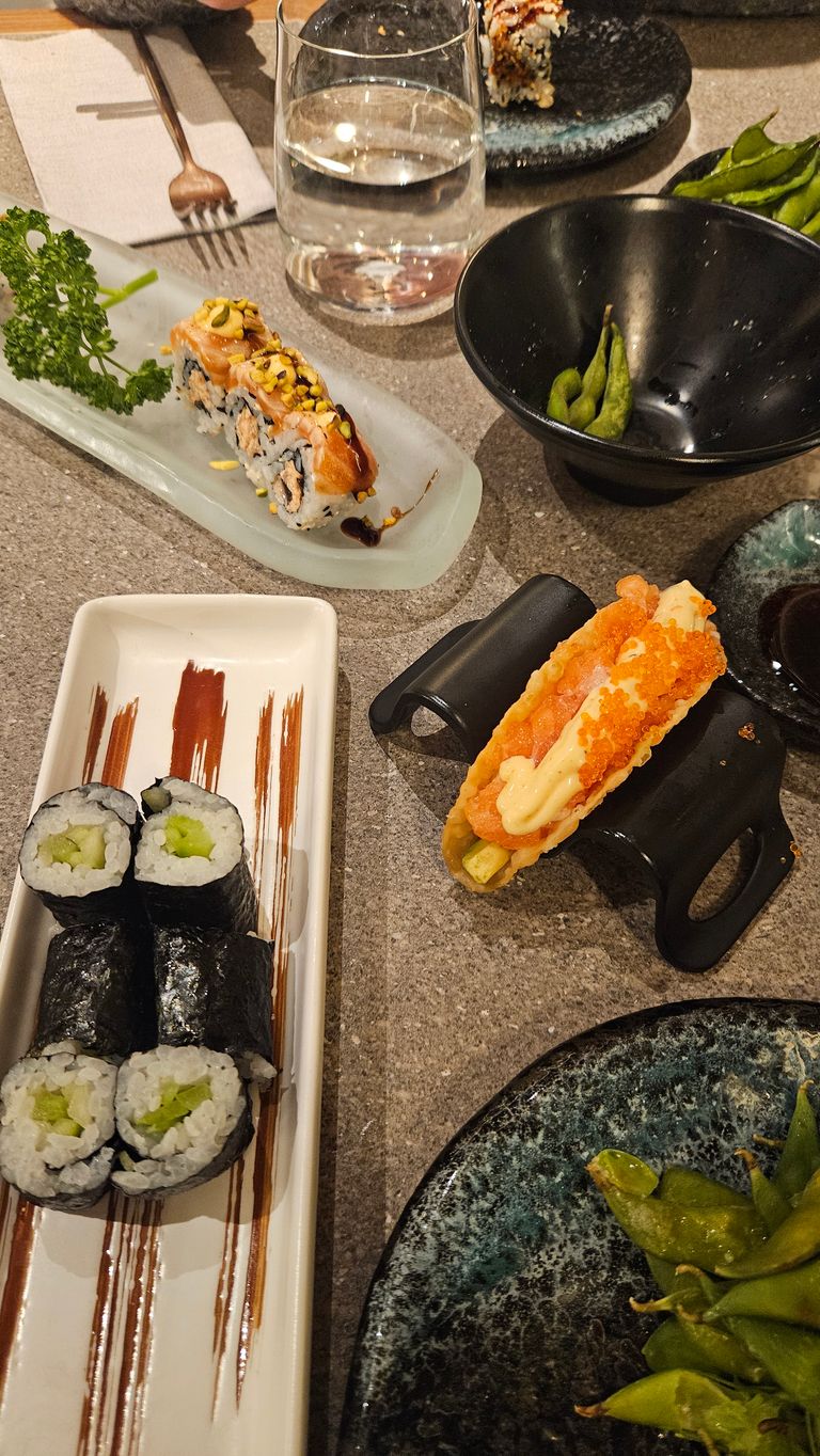 Various sushi rolls, edamame in a bowl, a glass of water, and a sushi taco on a gray table.