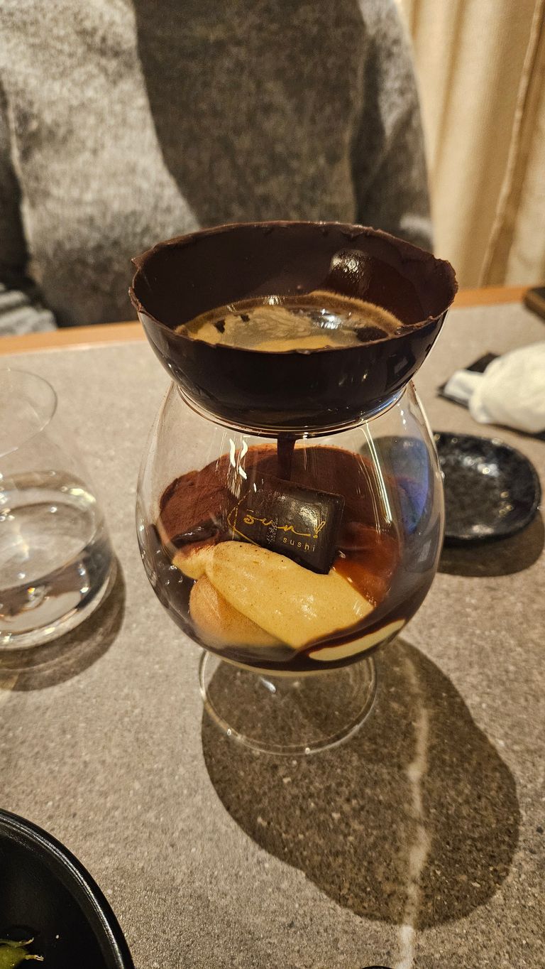 A dessert glass containing creamy pastry and cocoa layers, topped with a glass holding hot coffee, supported by a chocolate bowl, on a gray table with a glass of water and a person in the background.