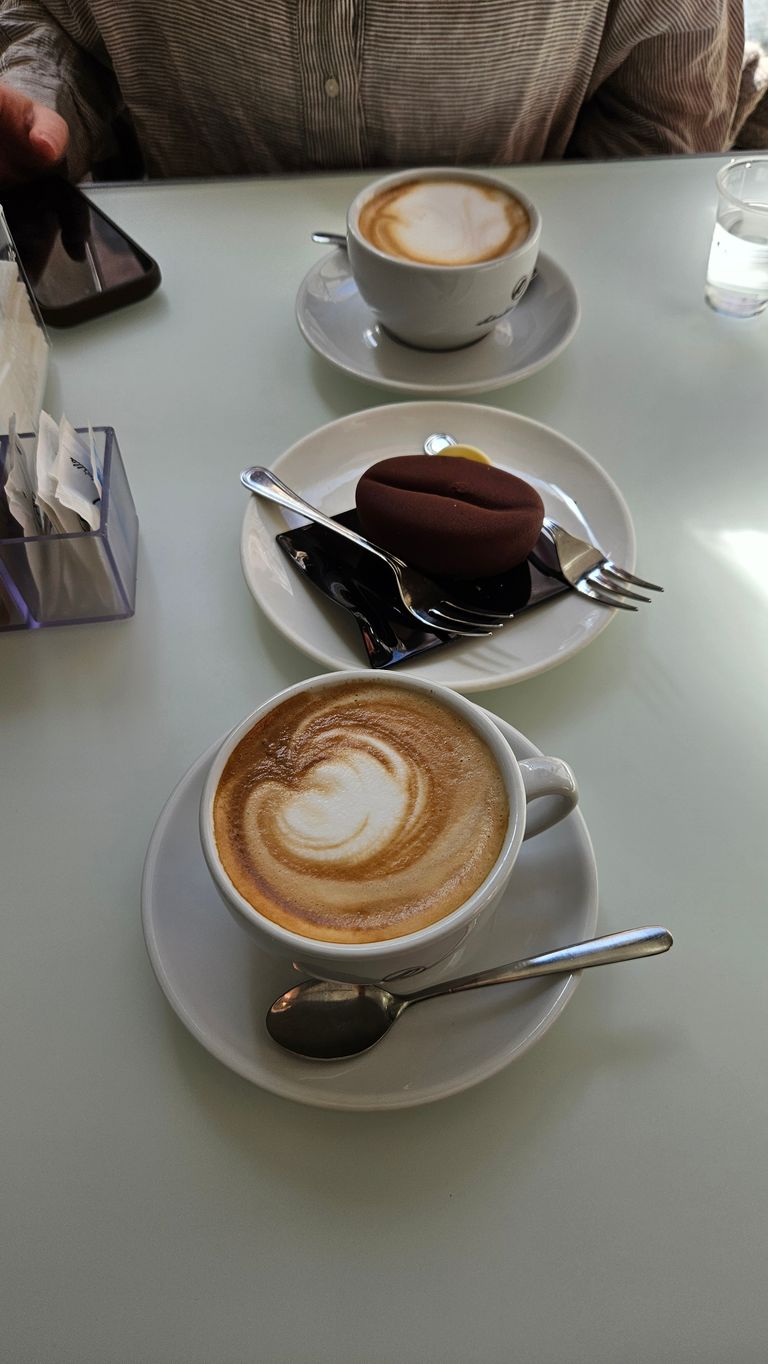 Two cups of cappuccino on a white table. Between the cups is a small plate with a coffee bean-shaped chocolate dessert and two small forks. A person in a striped shirt is sitting at the table holding a smartphone.