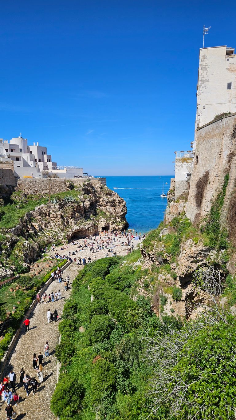View from above of a lively coastal town with whitewashed houses, pathways filled with people, and rocky cliffs leading to the blue sea.