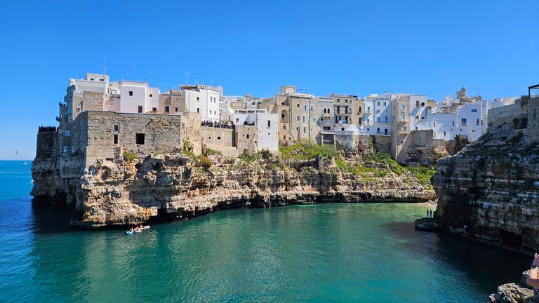Cliffside with white buildings on a rocky outcrop above clear turquoise sea under a clear blue sky.