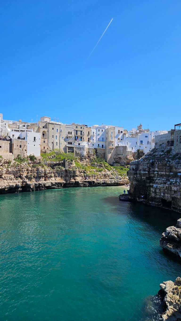 Coastal town with white and stone buildings on cliffs above clear blue-green water under a cloudless blue sky with a plane contrail.