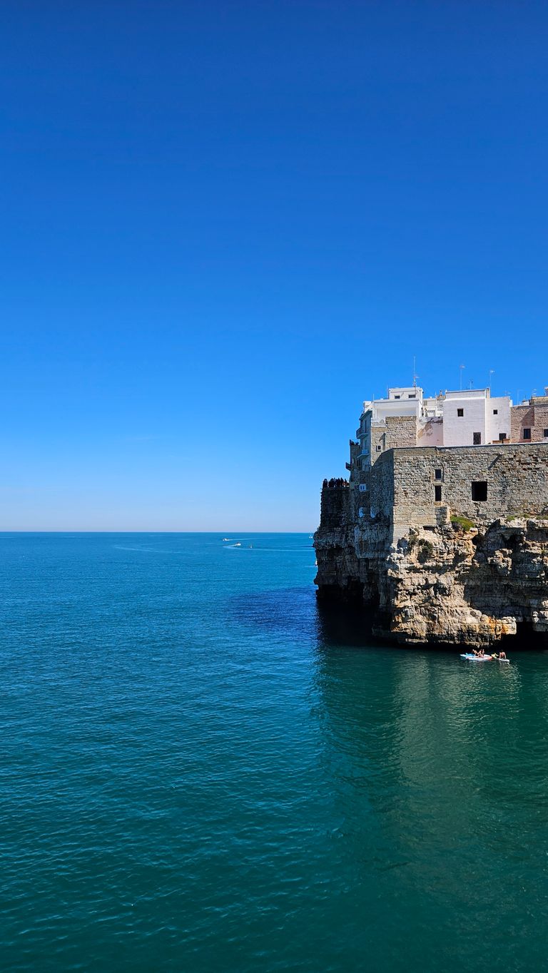 A stone coastal town with white houses perched on tall cliffs over a calm blue sea under a clear, cloudless sky.