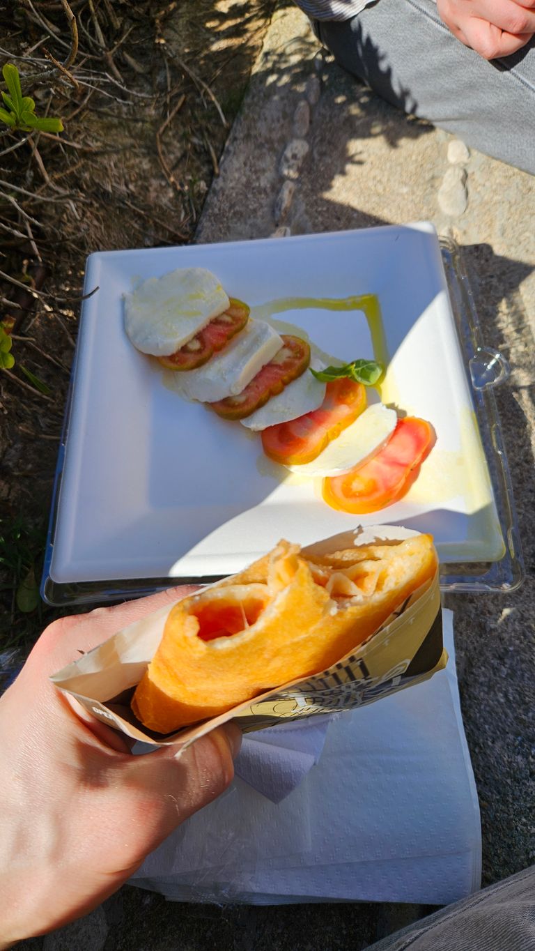 A hand holding a filled, sliced bread roll, in the background a plate with tomato slices and mozzarella, garnished with a basil leaf and olive oil, outdoors on a stone surface.