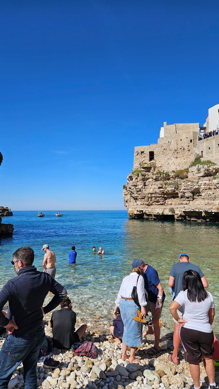 People on a pebble beach by clear blue sea water, some wading in, with an old stone building on a cliff in the background under a clear blue sky.