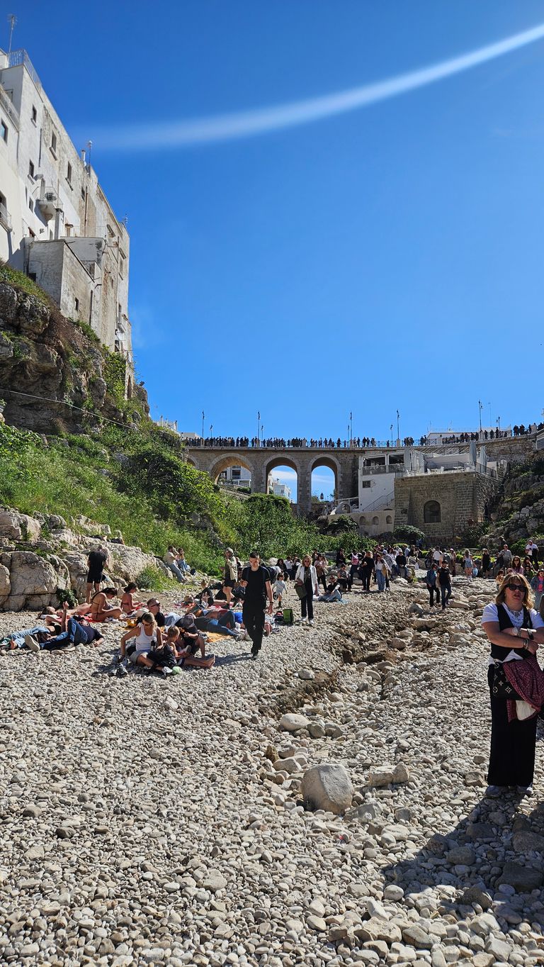 Many people relaxing and walking on a pebble-covered riverbed on a sunny day with green hills and historic bridges in the background.