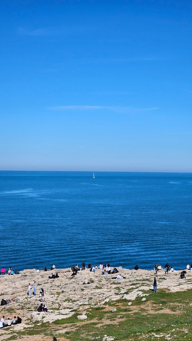 People sitting and standing on a rocky coastline overlooking the calm blue sea under a clear blue sky.