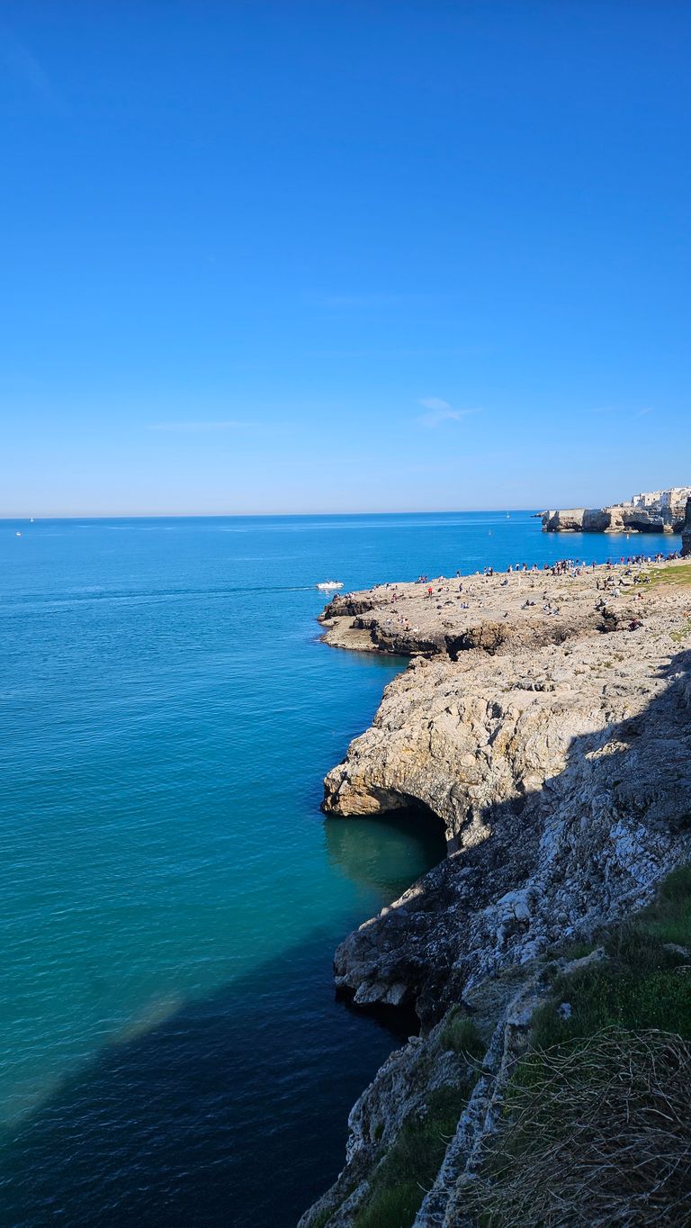 Rocky cliffs along the coast with clear blue sea and blue sky, people sitting on the rocks, a small boat sailing on the water.