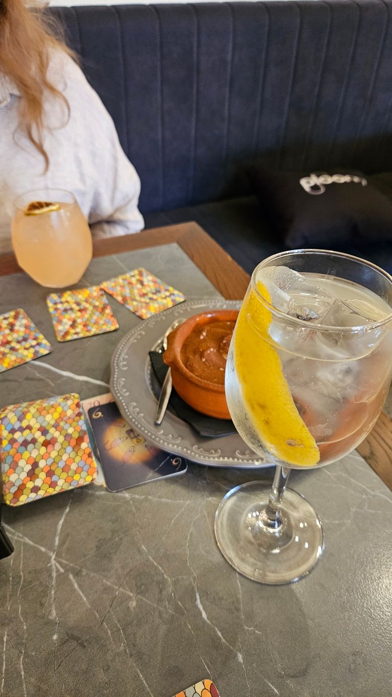 Drinks on a marble or stone table with patterned cards and a brown dessert in an earthenware pot, two people sitting comfortably on a padded bench.