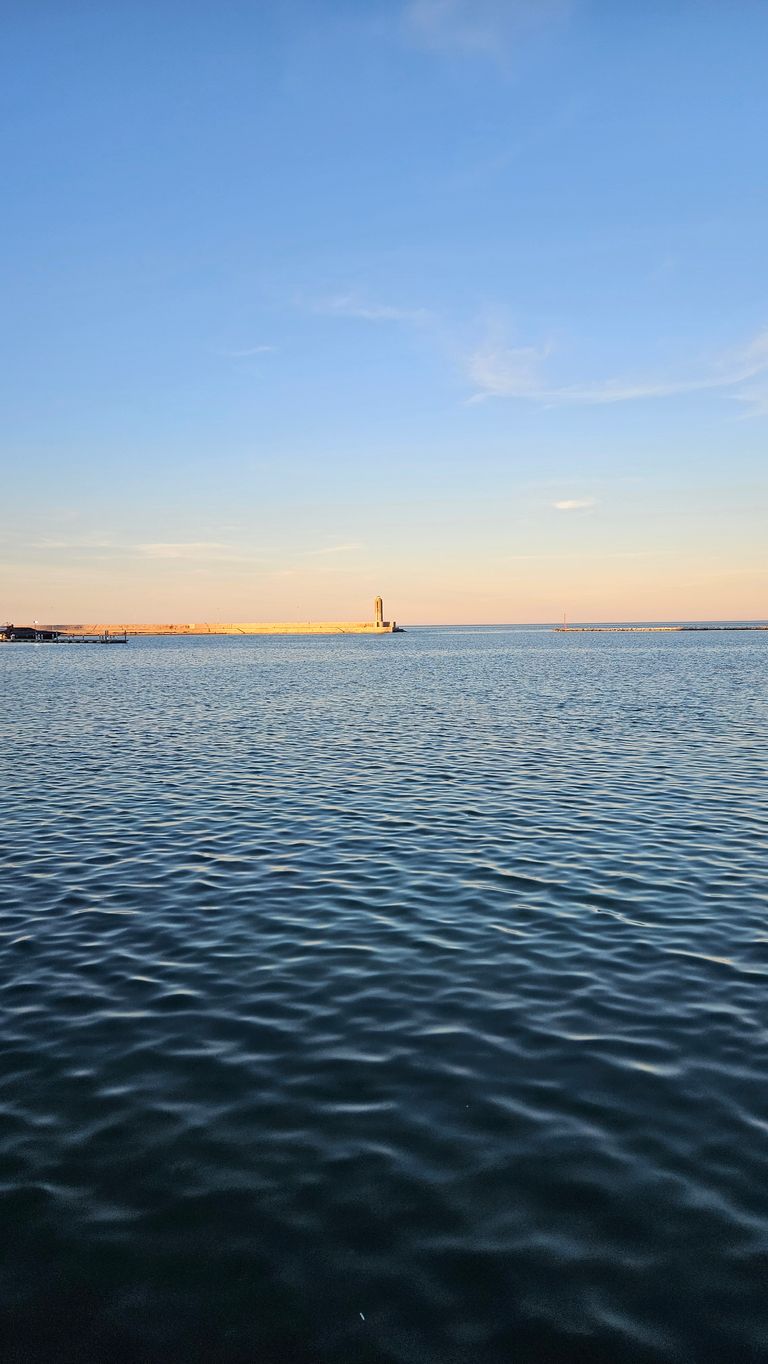 Calm sea or lake water at sunset with a clear sky and a small lighthouse on a pier visible on the horizon.