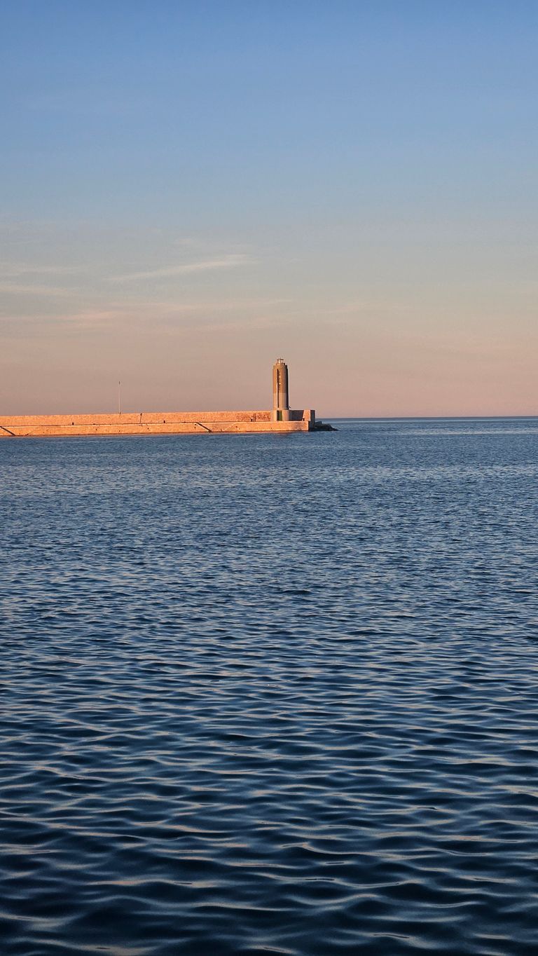 A long stone pier with a small lighthouse at the end extending over the calm sea under a clear sky.