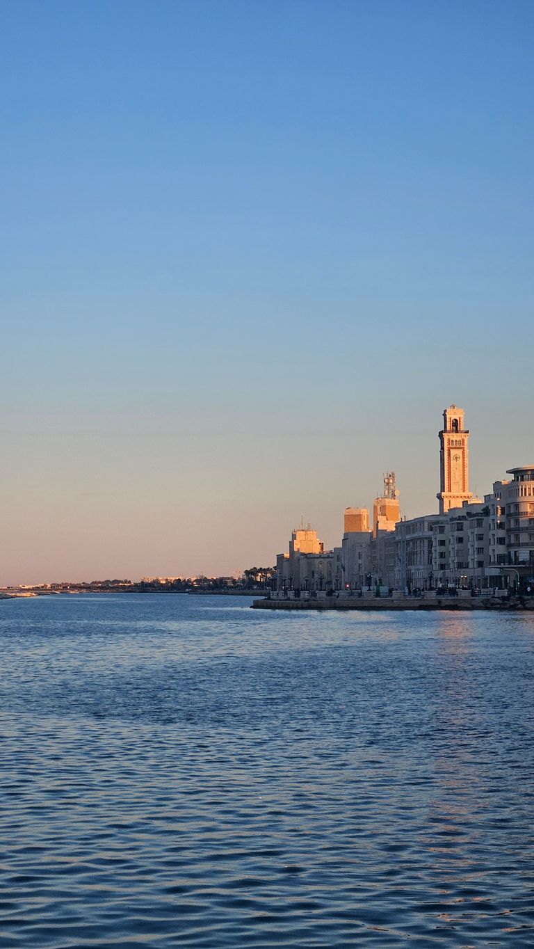 View of the waterfront promenade with historic buildings and a tower at sunset, calm water in the foreground.