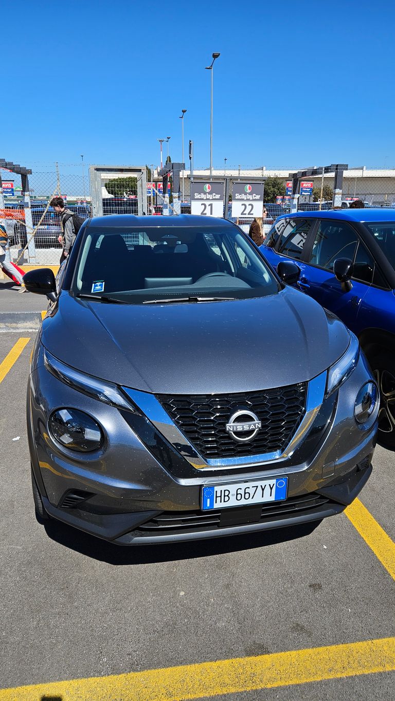 Gray Nissan SUV parked in a lot in front of a fence with "Sicily by Car" signs and parking spots 21 and 22 under clear blue sky.