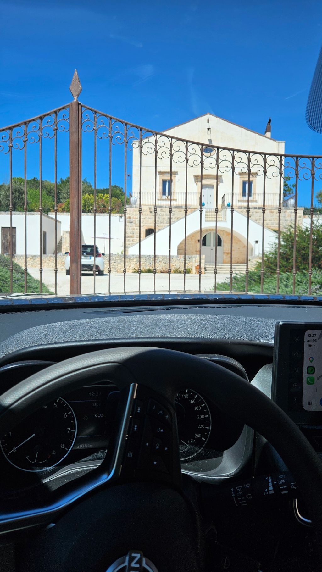 View from inside a car through a wrought iron garden gate towards a large white house with stairs and a blue sky.