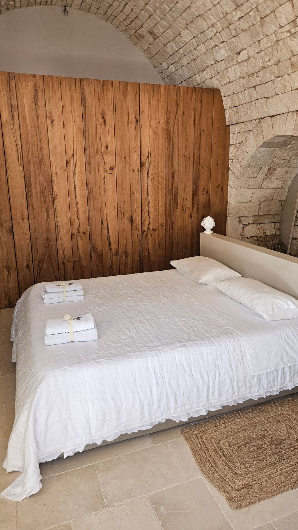 Simple bedroom with a bed covered in a white bedspread, two pillows, and two stacks of folded white towels. The wall is made of wooden panels next to a light stone arch.