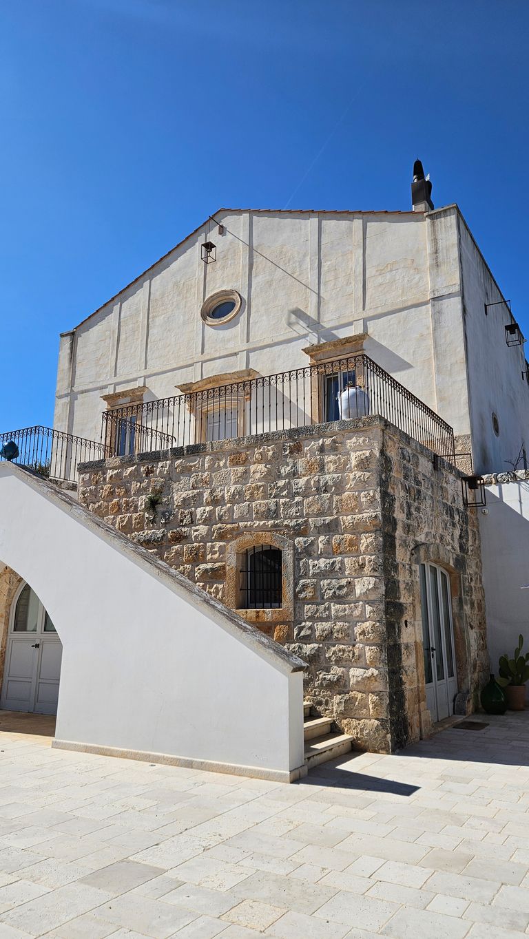 White two-story house with a sophisticated staircase, stone wall on the lower level, and clear blue sky in the background.