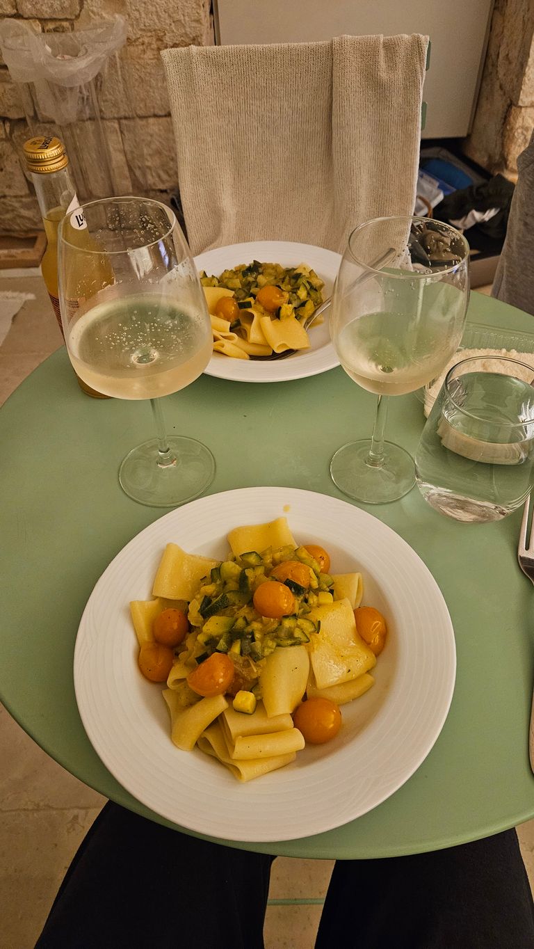 A green table with two plates of pasta topped with a sauce made of yellow cherry tomatoes and vegetables, accompanied by two glasses of white wine and a glass of water. A chair with a light-colored towel is visible in the background.