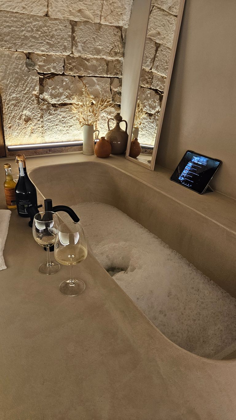 A built-in bathtub filled with foam, two wine glasses on the edge, and a mirror wall in front of a backlit stone wall. A ochre vase with dried flowers and a tablet on the bathtub ledge are also visible.