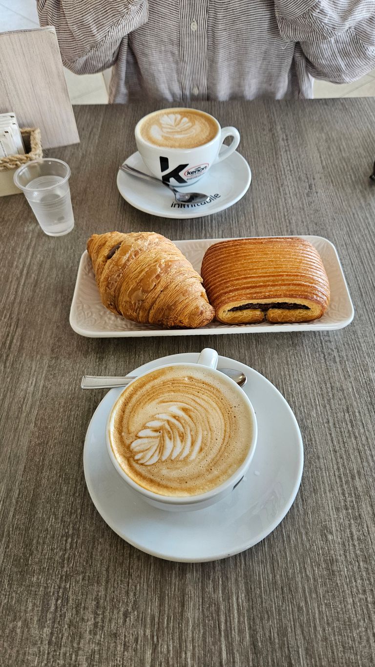 Two cups of coffee with milk foam art on a table, along with a rectangular white plate holding a croissant and a chocolate croissant, with a person wearing a striped shirt in the background.