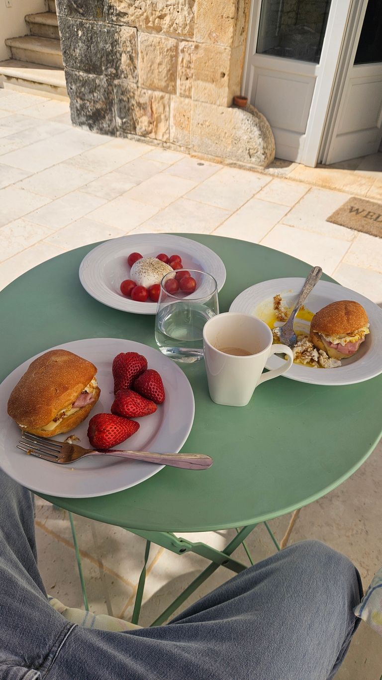 Green round table with breakfast items including a sandwich with strawberries on a plate, a white coffee cup, a glass of water, and a plate with tomatoes and cheese. Stone wall and stone stairs in the background.