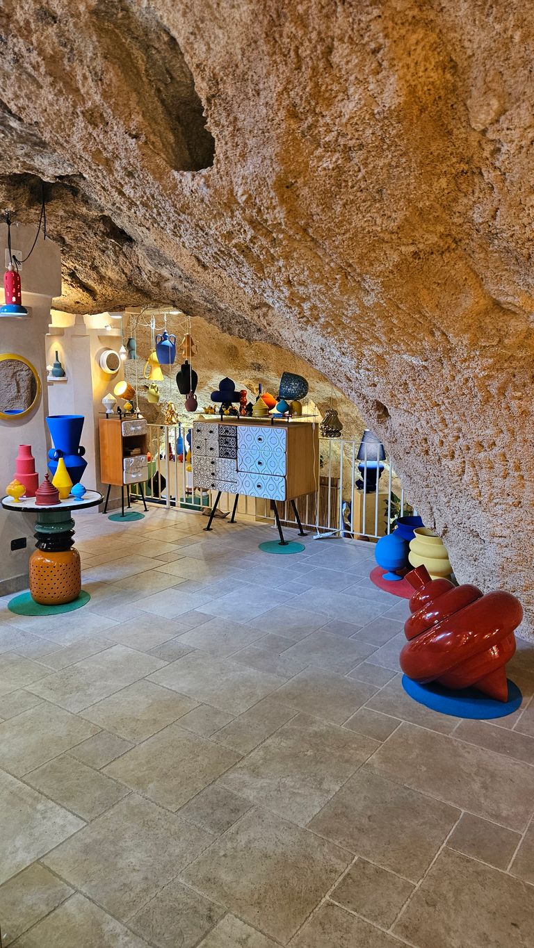 A bright interior space with a vaulted rough natural stone ceiling, decorated with colorful modern ceramic vases and sculptures placed on cabinets and pedestals.