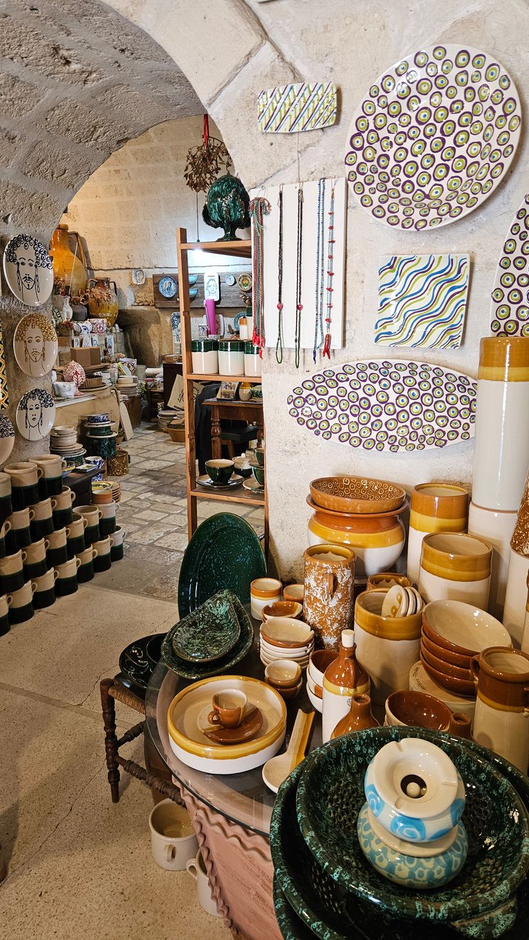Ceramic shop with a variety of pottery including plates, bowls, mugs, and handmade jewelry hanging on stone walls with an archway.