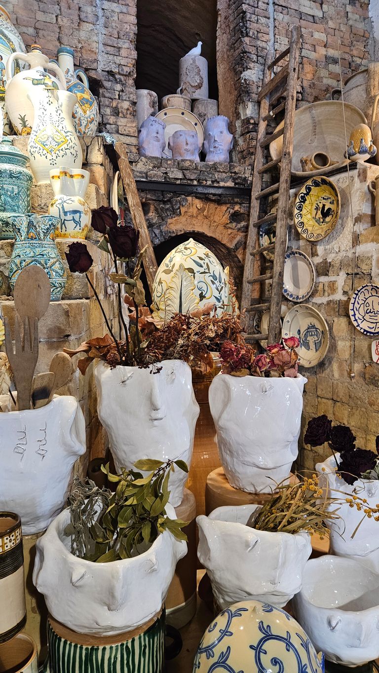 A collection of white ceramic vases with human facial features, filled with dried flowers and leaves, against a stone wall adorned with decorative ceramic plates and old ladders.