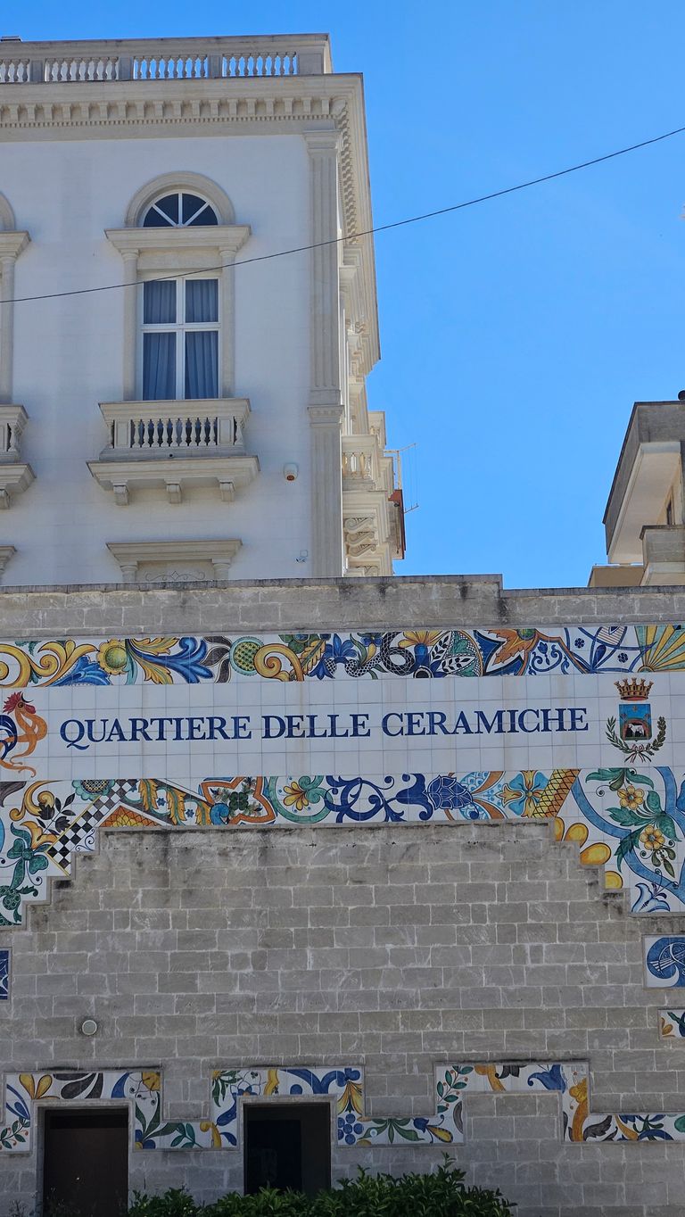 Exterior view of a building with a decorative colorful tiled wall displaying the text "QUARTIERE DELLE CERAMICHE" in blue letters. Above are two adjacent white buildings against a clear blue sky.