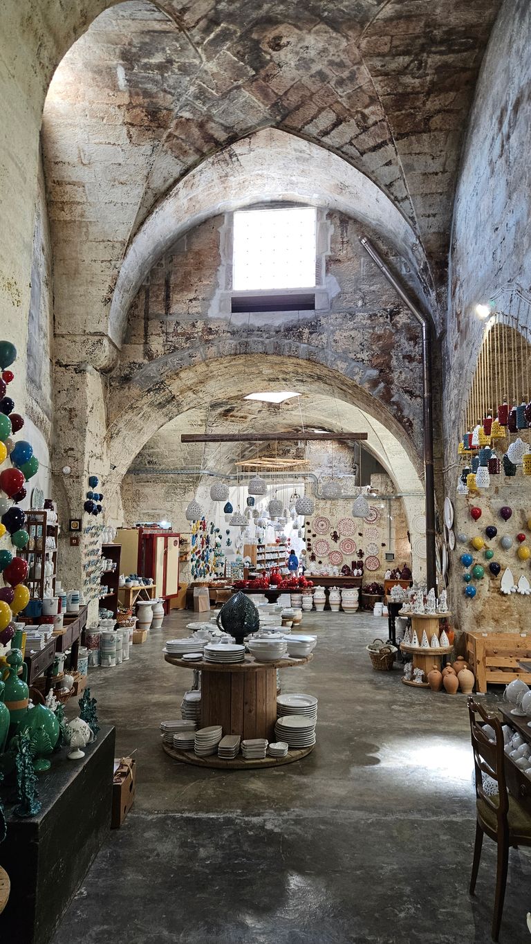 Interior of a stone vaulted building displaying colorful handicrafts and ceramics on tables and shelves.