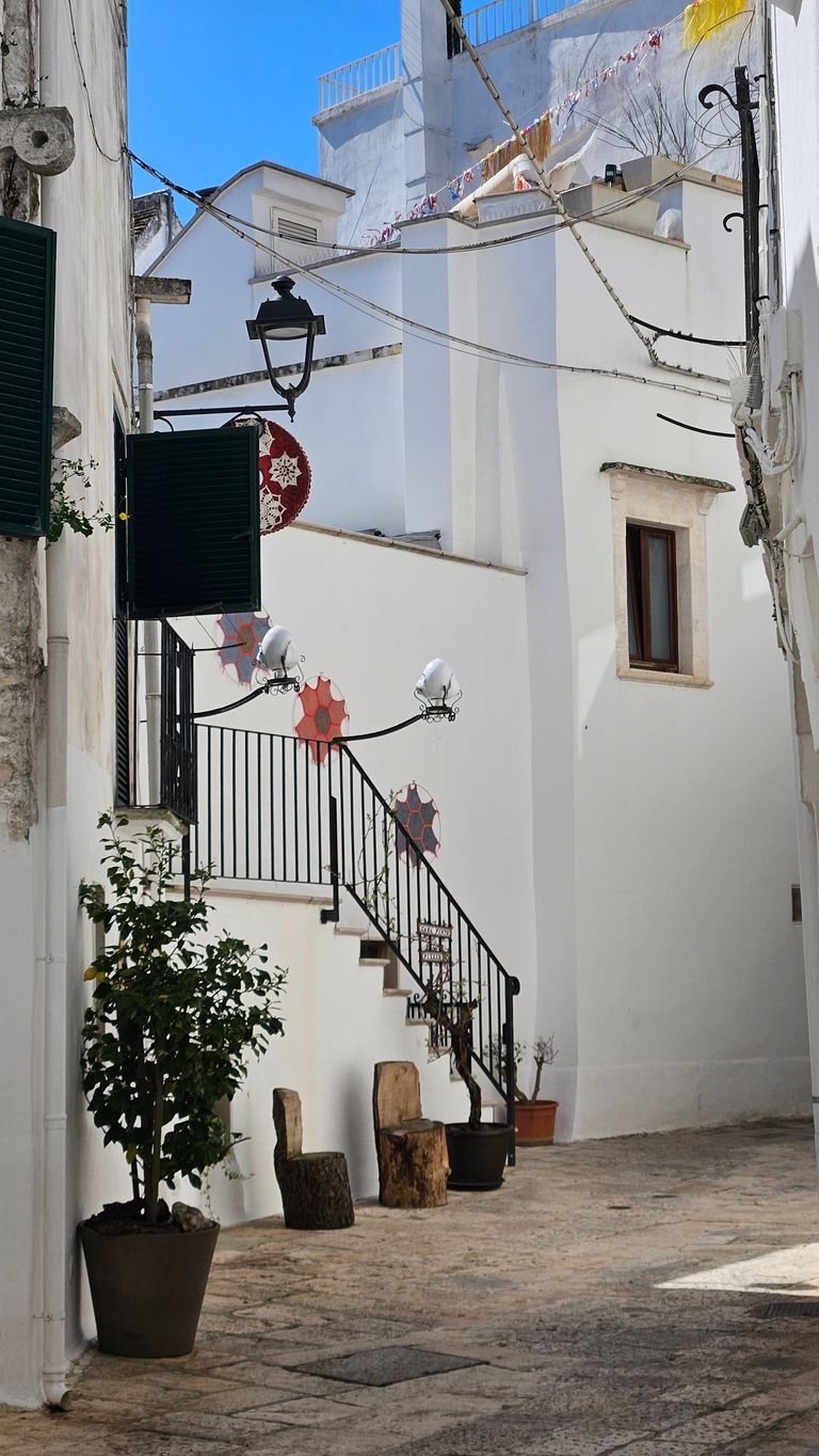 Narrow, bright alley with white houses, black railing on a staircase, potted plants, and colorful decorative elements on the wall and staircase.